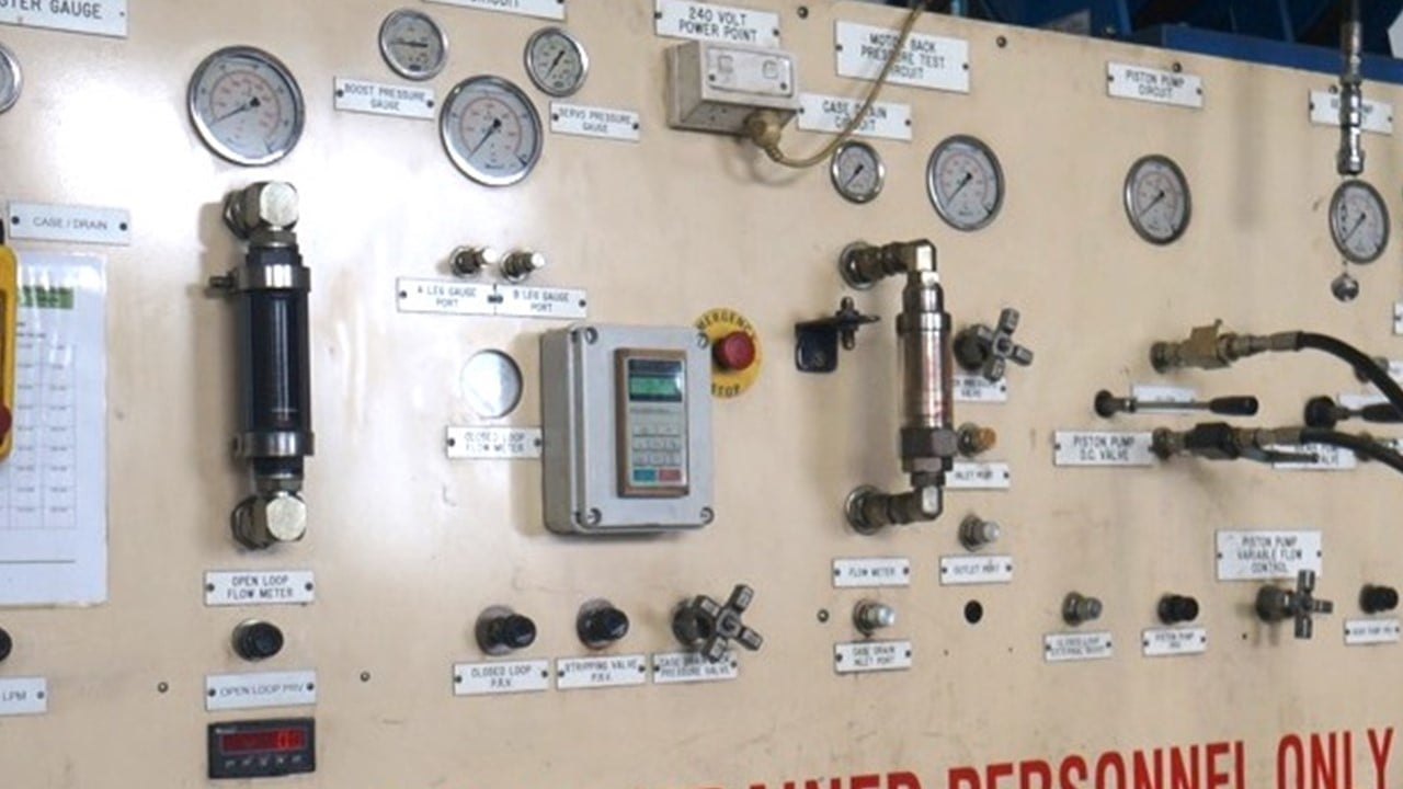 Industrial control panel with gauges, switches, and mechanical components mounted on a beige wall with labels